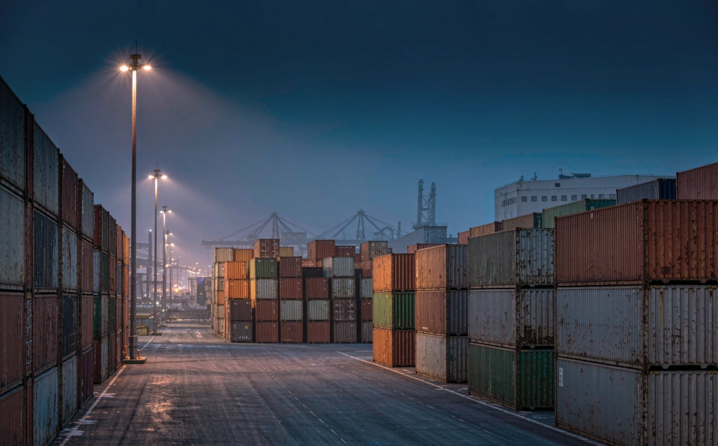 Container terminal at blue hour with warm orange floodlight pools between stacked freight containers